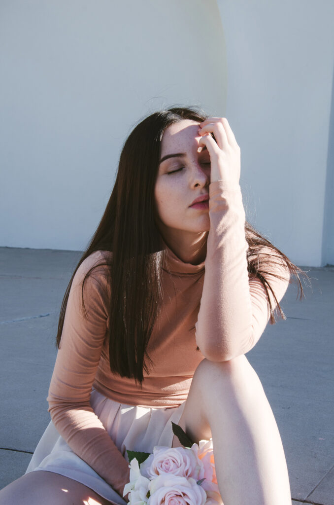 Gabriela Inzunza sits against a white wall her left hand touching her forehead while holding a bouquet of pink and white roses.